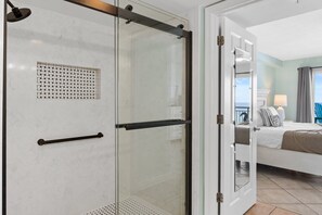 A hotel room with a glass shower enclosure, a bed with white bedding, and a view of the sea through the open door. - Pelican Isle 508 - Sandcastle Shores (Fort Walton Beach)
