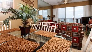 Un salón amplio con una mesa de comedor de cristal, sillas blancas, una planta grande en maceta, un sofá estampado y un ventilador de techo.