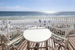 A balcony with a white table and two striped chairs, overlooking a beach with umbrellas and people. - Gulfside 503 - Tales From the Tide (Fort Walton Beach)