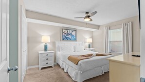 Un dormitorio con cama, mesitas de noche, ventilador de techo, un cuadro y una ventana con cortinas.