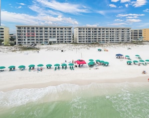 A beach with multiple green umbrellas and lounge chairs, a building with balconies, and a sandy shoreline. - Gulf Dunes 416 - Ocean's Edge (Fort Walton Beach)