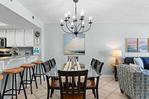 A dining area with a glass table, wooden chairs, a chandelier, and a painting on the wall. - Gulf Dunes 416 - Ocean's Edge (Fort Walton Beach)