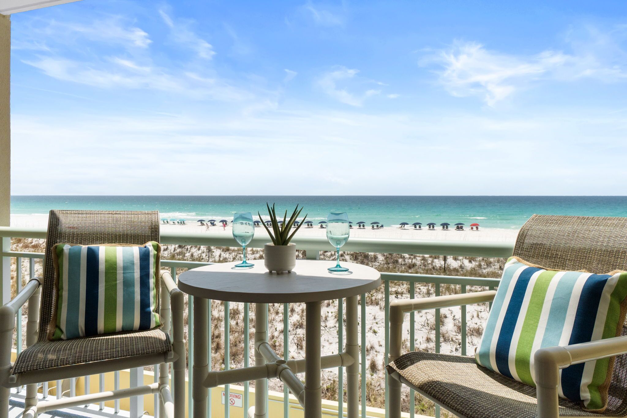 Un balcon avec des fauteuils en osier et une petite table, donnant sur une plage animée et un ciel dégagé.