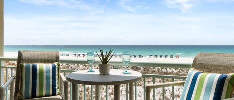 A balcony with wicker chairs and a small table, overlooking a beach with people and a clear sky.