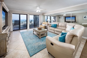 A spacious living room with a large sofa, a coffee table, and a television. There is a balcony with a view of the ocean. - Surf Dweller 502 - The Dune Retreat (Fort Walton Beach)