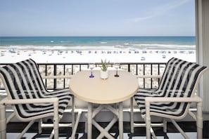 A balcony with two striped chairs and a small table, overlooking a beach with umbrellas and people. - Surf Dweller 502 - The Dune Retreat (Fort Walton Beach)