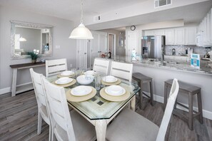 A modern kitchen with white cabinets, a stainless steel refrigerator, and a glass dining table set for six. - Gulf Dunes 401 - Ocean Eyes (Fort Walton Beach)
