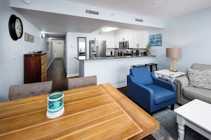 A modern kitchen with white cabinets, a microwave, and a refrigerator. There is a dining area with a wooden table and chairs, and a living room with a blue sofa and a gray couch. - Pelican Isle 510 - Sunset Serenade (Fort Walton Beach)