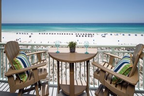 En balkon med to træstole og et bord, der giver udsigt over en strand med parasoller og mennesker.