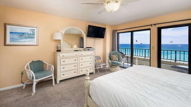A bedroom with a bed, dresser, chairs, a TV, and a balcony with a view of the ocean. - Surf Dweller 603 - Swaying Palms (Fort Walton Beach)