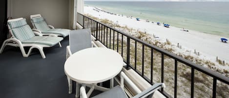 A balcony with two lounge chairs and a small table, overlooking a beach with umbrellas and people.