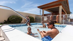 A swimming pool area with a diving board, a covered seating area, and a building with a red roof. - Surf Dweller 603 - Swaying Palms (Fort Walton Beach)