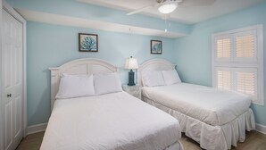 A bedroom with two beds, a ceiling fan, and a window with shutters. - Gulf Dunes 301 - Seas the Day (Fort Walton Beach)