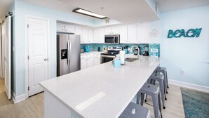 A modern kitchen with white cabinets, a large island, and a blue accent wall. - Gulf Dunes 301 - Seas the Day (Fort Walton Beach)