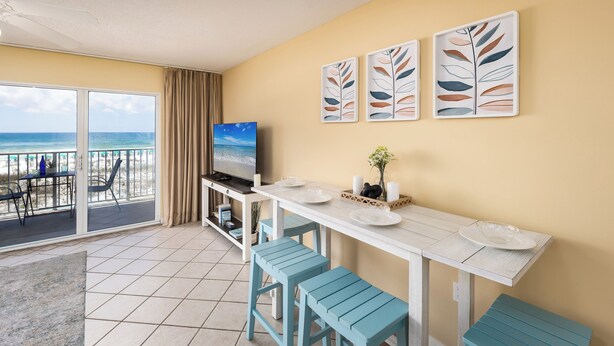 A room with a balcony overlooking the beach, a TV, a dining table with chairs, and artwork on the wall. - Gulf Dunes 207 - Blue Skies & Beach Vibes (Fort Walton Beach)