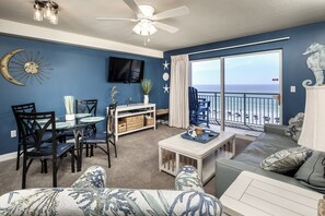 Ein Wohnzimmer mit Essbereich, Deckenventilator, Wand-Fernseher und großem Fenster mit Blick auf den Strand.