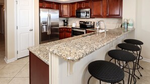 A kitchen with a granite countertop, stainless steel appliances, wooden cabinets, and four black bar stools. - Gulf Dunes 315 - Salt and Serenity (Fort Walton Beach)