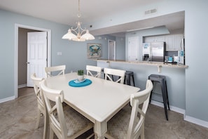 A dining area with a white table and chairs, a blue placemat, and a hanging light fixture. - Gulf Dunes 412 - Sunset Fiesta (Fort Walton Beach)