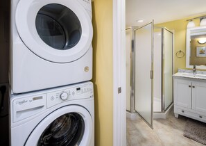 A laundry area with stacked washers and dryers, a glass shower door, and a bathroom with a sink and mirror. - Pelican Isle 406 - Sunset Shores (Fort Walton Beach)