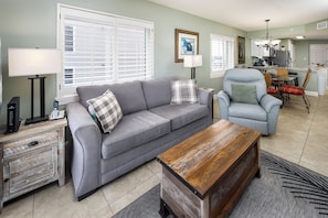 A living room with a grey sofa, a wooden coffee table, and a dining area with a chandelier. - Summerlin 305 - Breezy Palms (Fort Walton Beach)