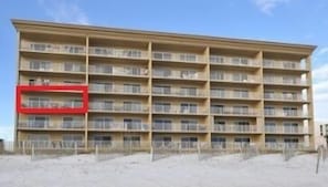 Een hotel met meerdere verdiepingen, balkons, een raam met rode omlijsting aan de linkerkant en een zandachtige ondergrond.