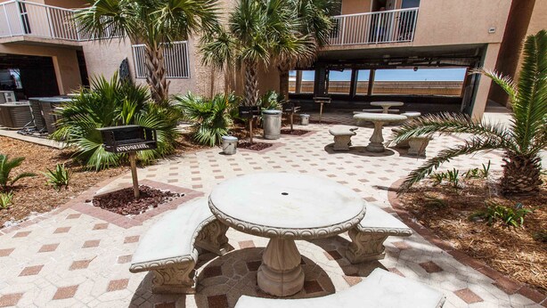 A courtyard with a stone table and benches, surrounded by palm trees and a building with a balcony. - Islander 3004 - Coral Dreams (Fort Walton Beach)