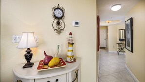 A hallway with a white console table featuring a lamp, a fruit bowl, and a lighthouse decoration. A wall clock and a thermostat are also visible. - Islander 3004 - Coral Dreams (Fort Walton Beach)