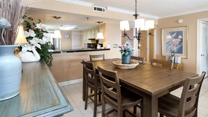 A dining area with a wooden table and chairs, a vase with flowers, and a kitchen in the background. - Surf Dweller 402 - Seashell Haven (Fort Walton Beach)