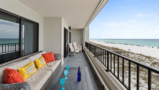 A balcony with a view of the beach, a sofa with cushions, and a table with glasses and a bottle. - Surf Dweller 402 - Seashell Haven (Fort Walton Beach)