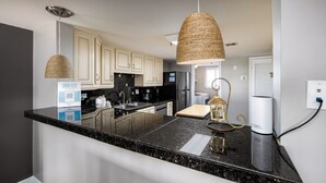 A kitchen with a granite countertop, white cabinets, a stainless steel refrigerator, and a hanging woven light fixture. - Surf Dweller 402 - Seashell Haven (Fort Walton Beach)