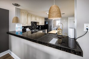 A kitchen with a granite countertop, white cabinets, a stainless steel refrigerator, and a hanging woven light fixture. - Surf Dweller 402 - Seashell Haven (Fort Walton Beach)