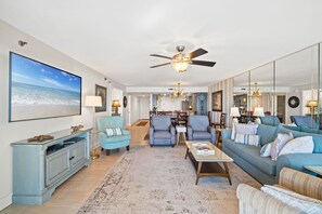 A spacious living room with a large TV, comfortable seating, a ceiling fan, and a view of the kitchen in the background. - Surf Dweller 205 - Seaspray Getaway (Fort Walton Beach)