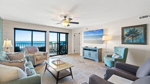 Ein geräumiges Wohnzimmer mit großem Fenster zum Meer, einem Deckenventilator und Blick auf den Strand.