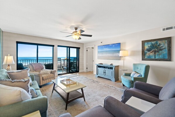 A spacious living room with a large window overlooking the ocean, a ceiling fan, and a view of the beach. - Surf Dweller 205 - Seaspray Getaway (Fort Walton Beach)