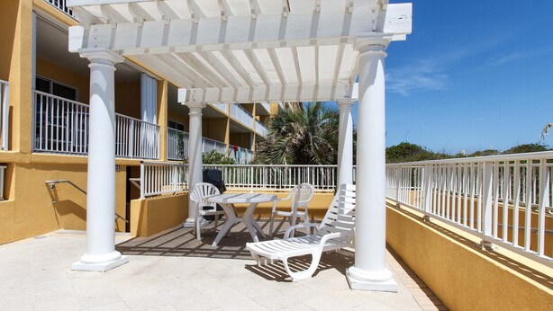 A covered outdoor seating area with white chairs and a table, surrounded by a white railing, under a pergola. - Gulf Dunes 402 - Palm Haven (Fort Walton Beach)