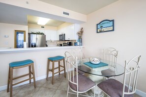 Un coin repas avec une table en verre et des chaises en métal blanc, une cuisine avec des meubles blancs et des appareils électroménagers, et un panneau décoratif sur le mur sur le thème de la plage.