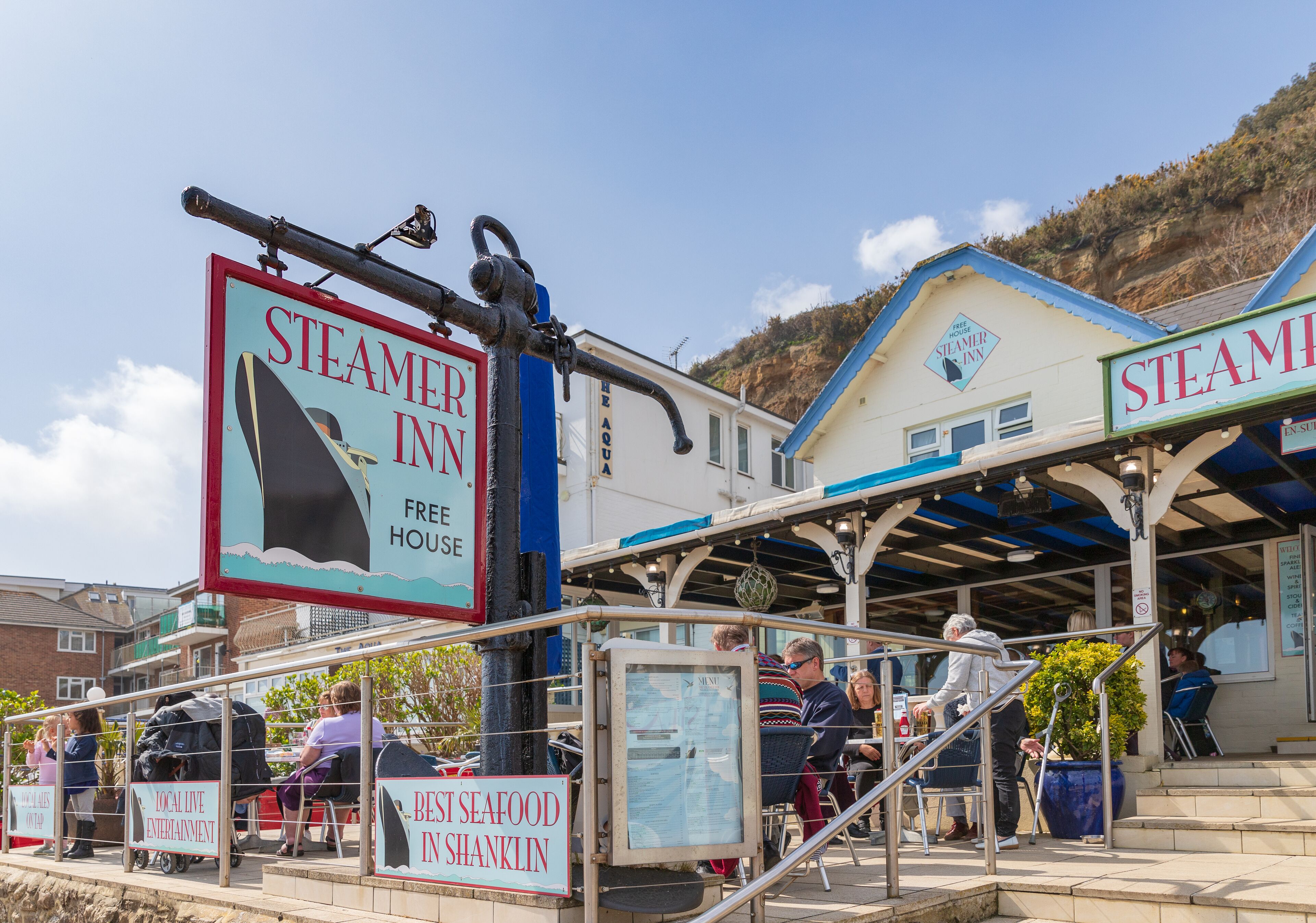 Ein Restaurant am Meer mit Außenbereich, einem Schild mit der Aufschrift 'STEAMER INN' und 'FREE HOUSE' sowie Blick aufs Meer.