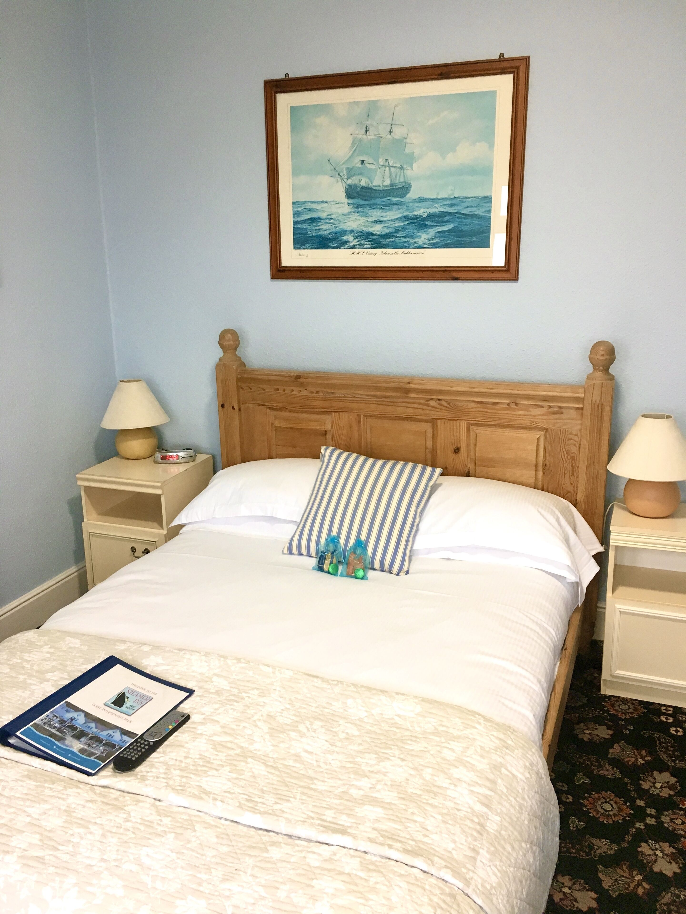 A neatly made bed with a striped pillow, a framed painting of a ship on the wall, and a remote control on the bed.
