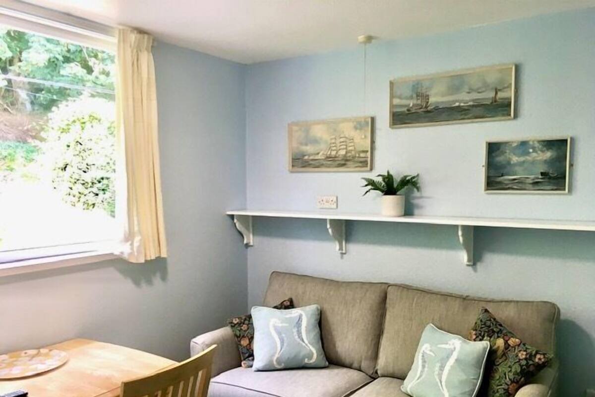 A room with a beige sofa, a wooden table, and a window with curtains. There are two wall-mounted shelves with paintings and a small potted plant.
