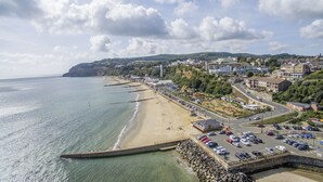 A coastal town with a sandy beach, a promenade lined with parked cars, and buildings along the hillside.