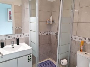 Et badeværelse med glasvægs brusekabine, en hvid vask med sæbedispenser og et toilet med toiletpapir.