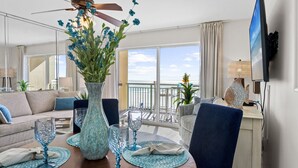 Een moderne woonkamer met een groot raam met uitzicht op de oceaan, een plafondventilator, een eettafel gedekt met blauw glaswerk, en een flatscreen televisie aan de muur.
