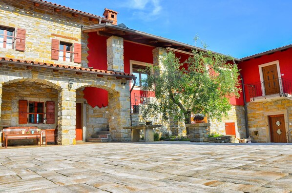 A courtyard with stone flooring, a wooden bench, and a building with red and yellow walls and stone pillars. - Apartament "Pomigranaj" for 4 person (Marezige)