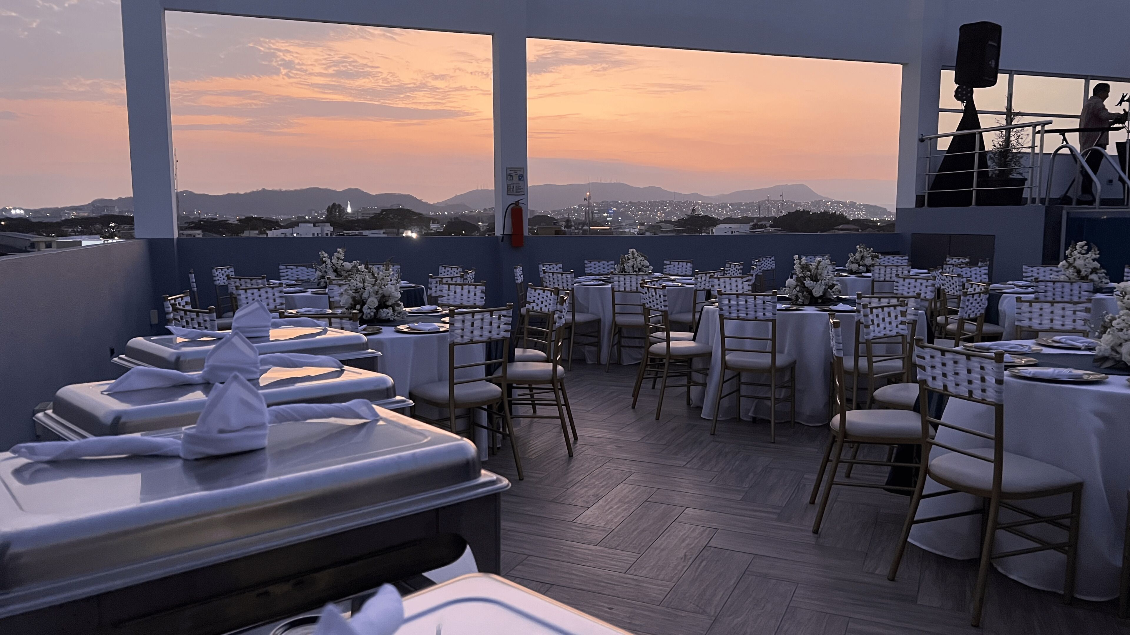 Un restaurante en la azotea con mesas dispuestas para comer, con vistas a un paisaje urbano al atardecer.