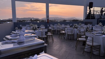 Un restaurante en la azotea con mesas dispuestas para comer, con vistas a un paisaje urbano al atardecer.