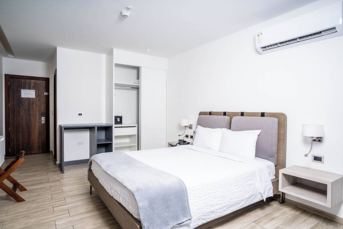 Una habitació d'hotel moderna amb llit, taula de nit, armari, escriptori i aire condicionat integrat.