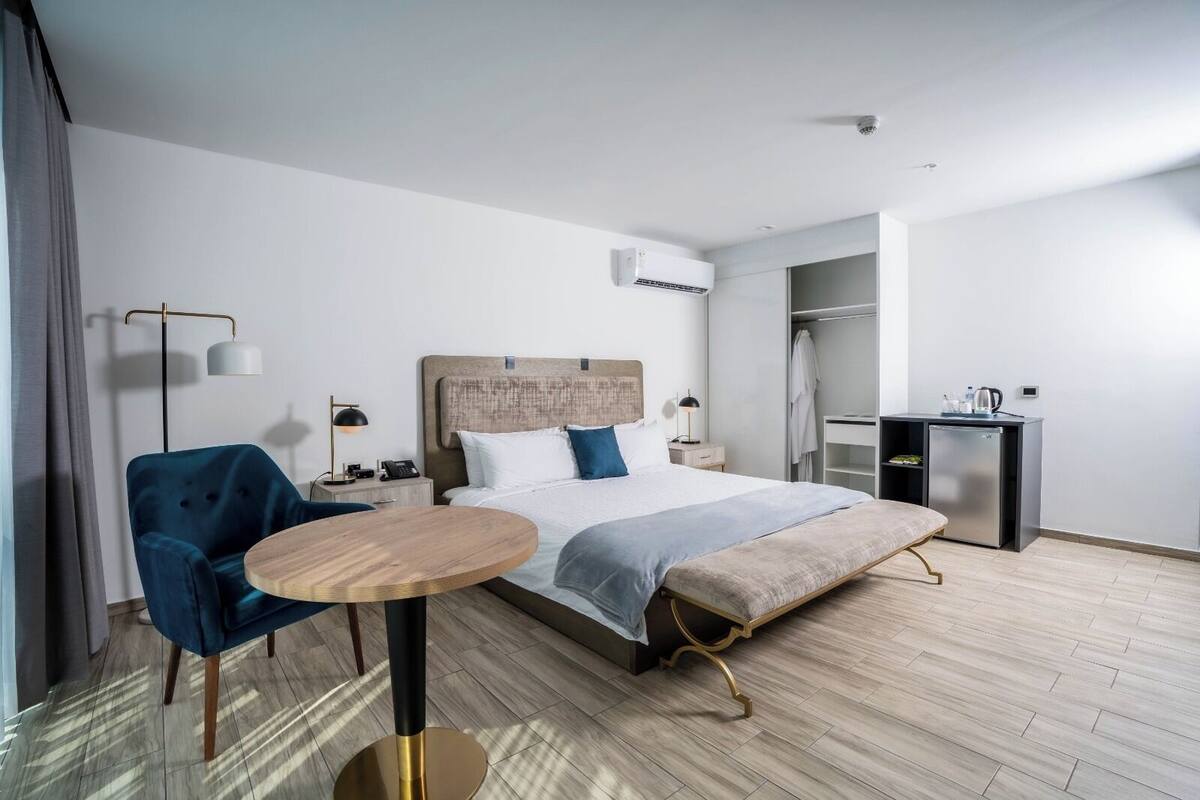 Habitació d'hotel moderna amb un llit gran, una taula rodona, un butac armchair blau, un escriptori amb nevera i un armari.