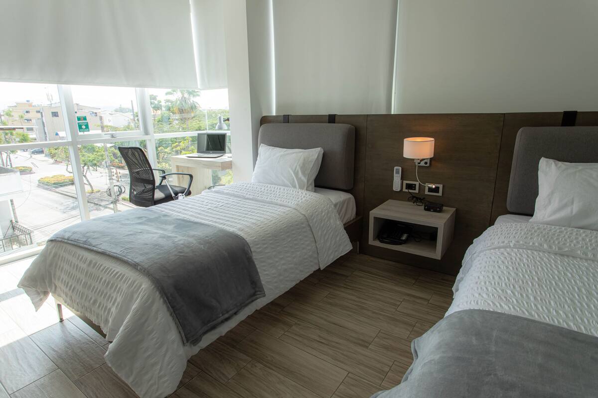 Habitació d'hotel amb dos llits, un escriptori, una cadira i una finestra que dona a edificis i vegetació.