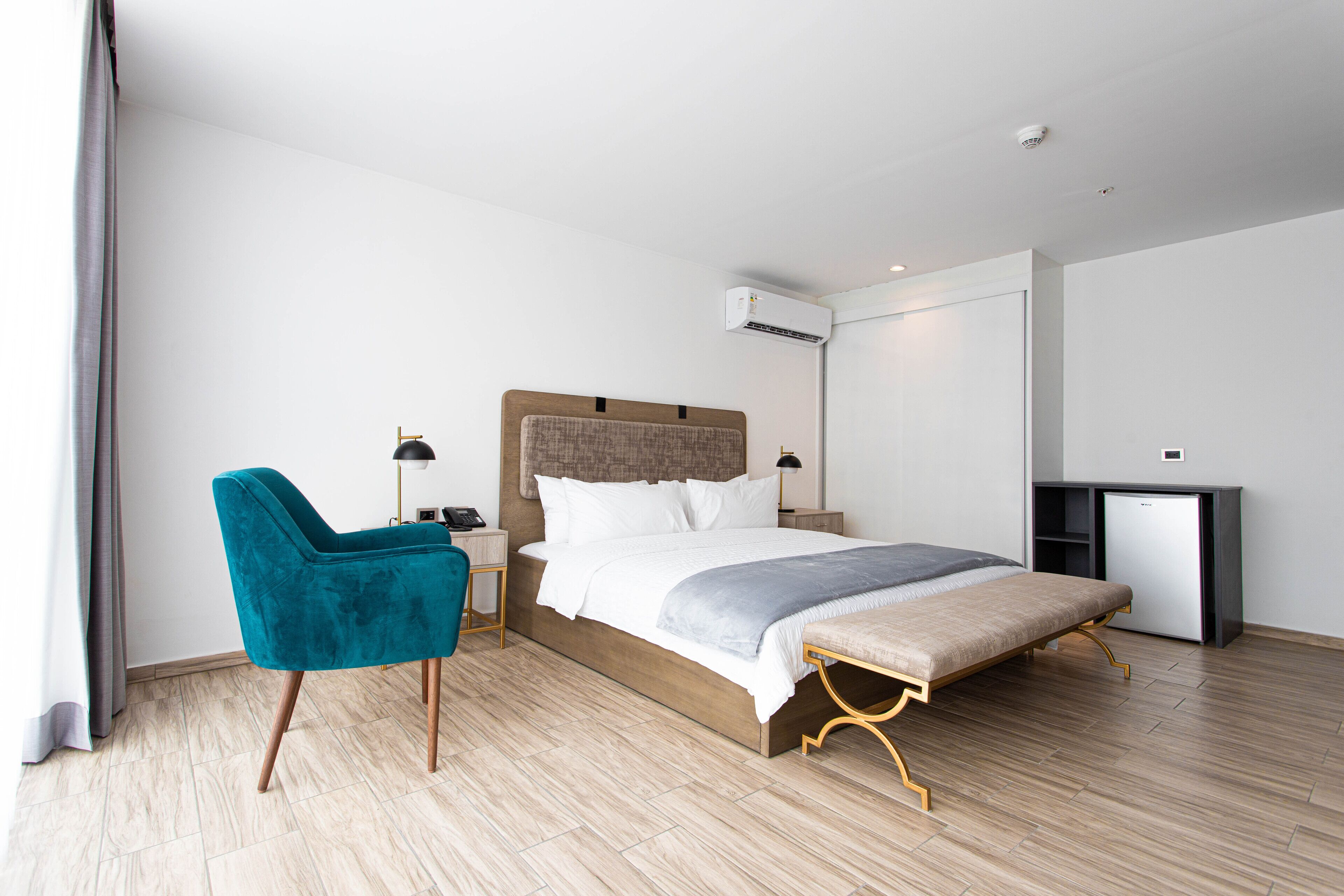 Ein modernes Hotelzimmer mit einem hölzernen Bett, einem blaugrünen Sessel, einem Schreibtisch und einem kleinen Kühlschrank.