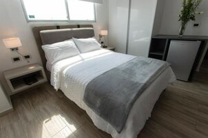 Un dormitorio moderno con cama, mesitas de noche, un escritorio y un refrigerador pequeño.